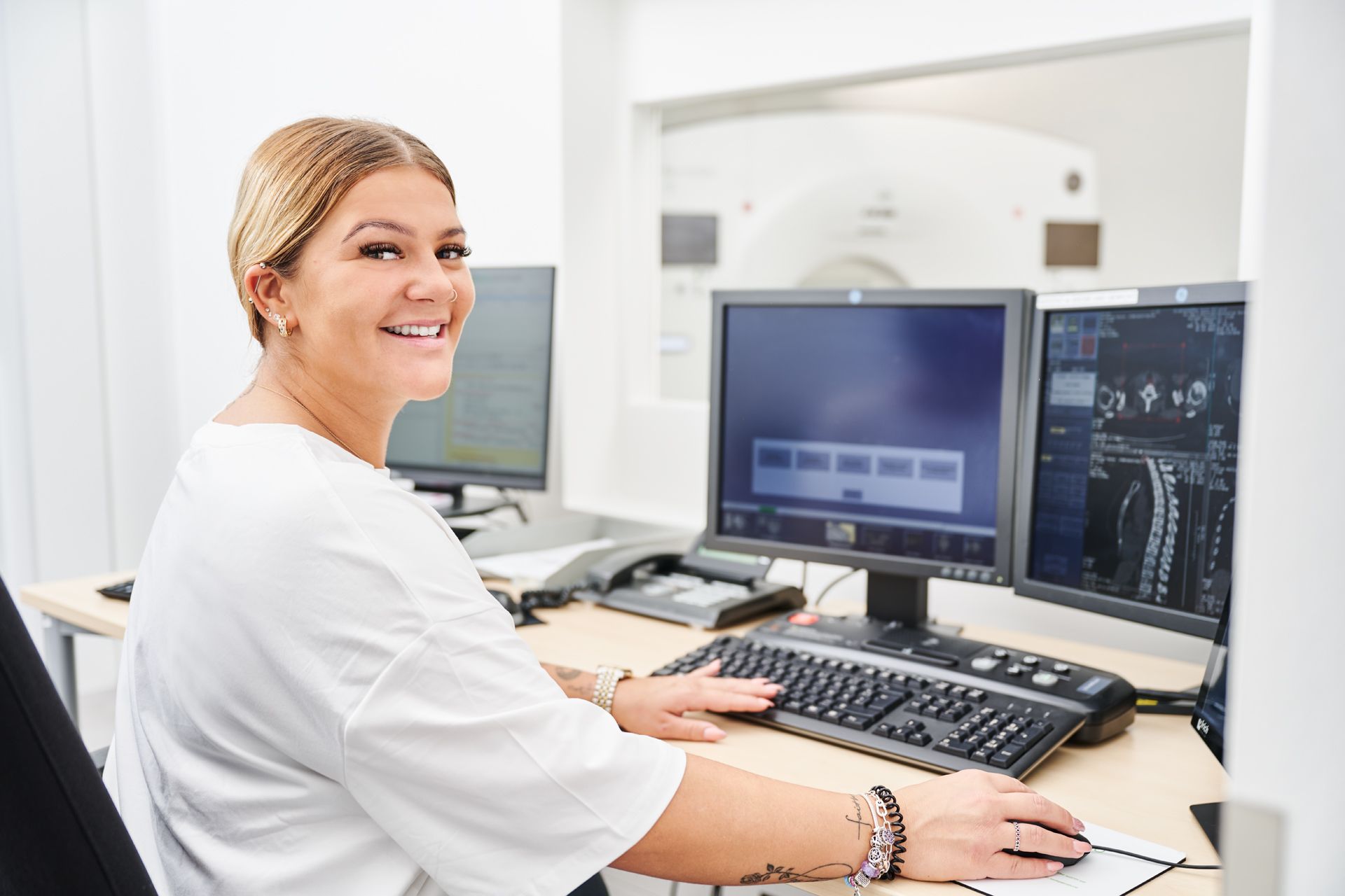 Medizinische Fachkraft sitzt vor einem Bildschirm mit CT-Aufnahmen und lächelt in die Kamera. Computer Bildschirm an.