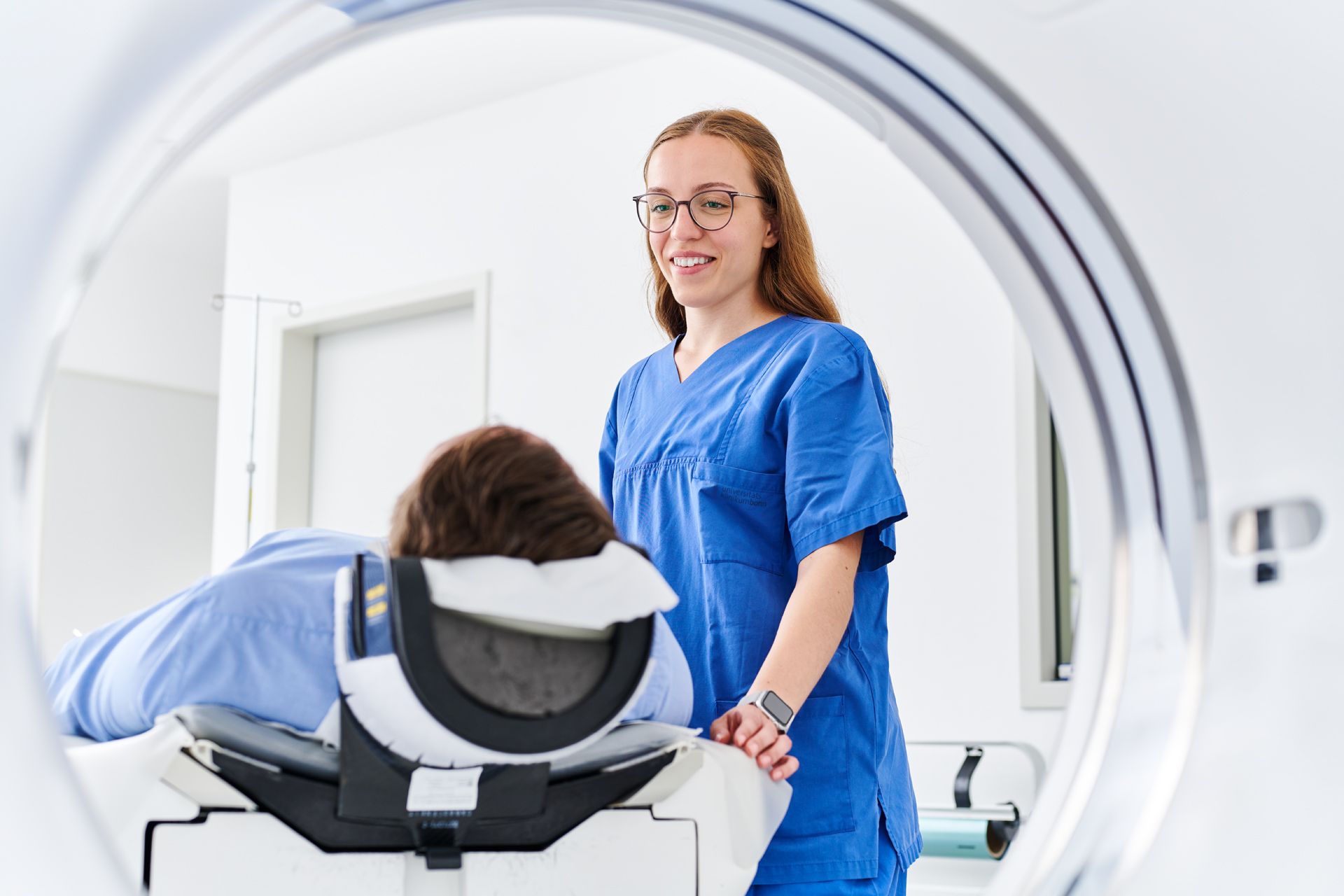 Blick von hinten durch eine CT-Röhre. Der Patient liegt in einem CT und wird von Medizinischer Fachkraft betreut. 