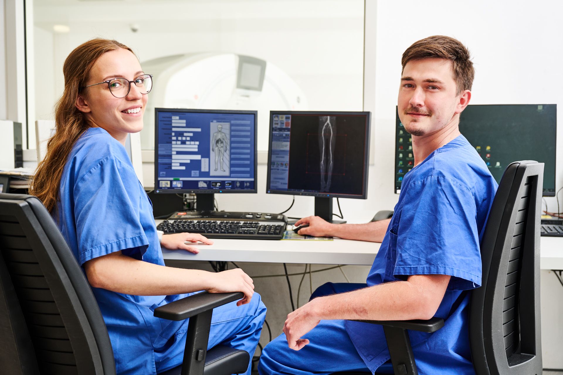 Zwei Medizinische Fachkräfte sitzen an einem Schreibtisch mit Conputerbildschirmen, auf denen CT-Bilder zu sehen sind. Sie schauen in die Kamera. einem Computer Bildschirm an.